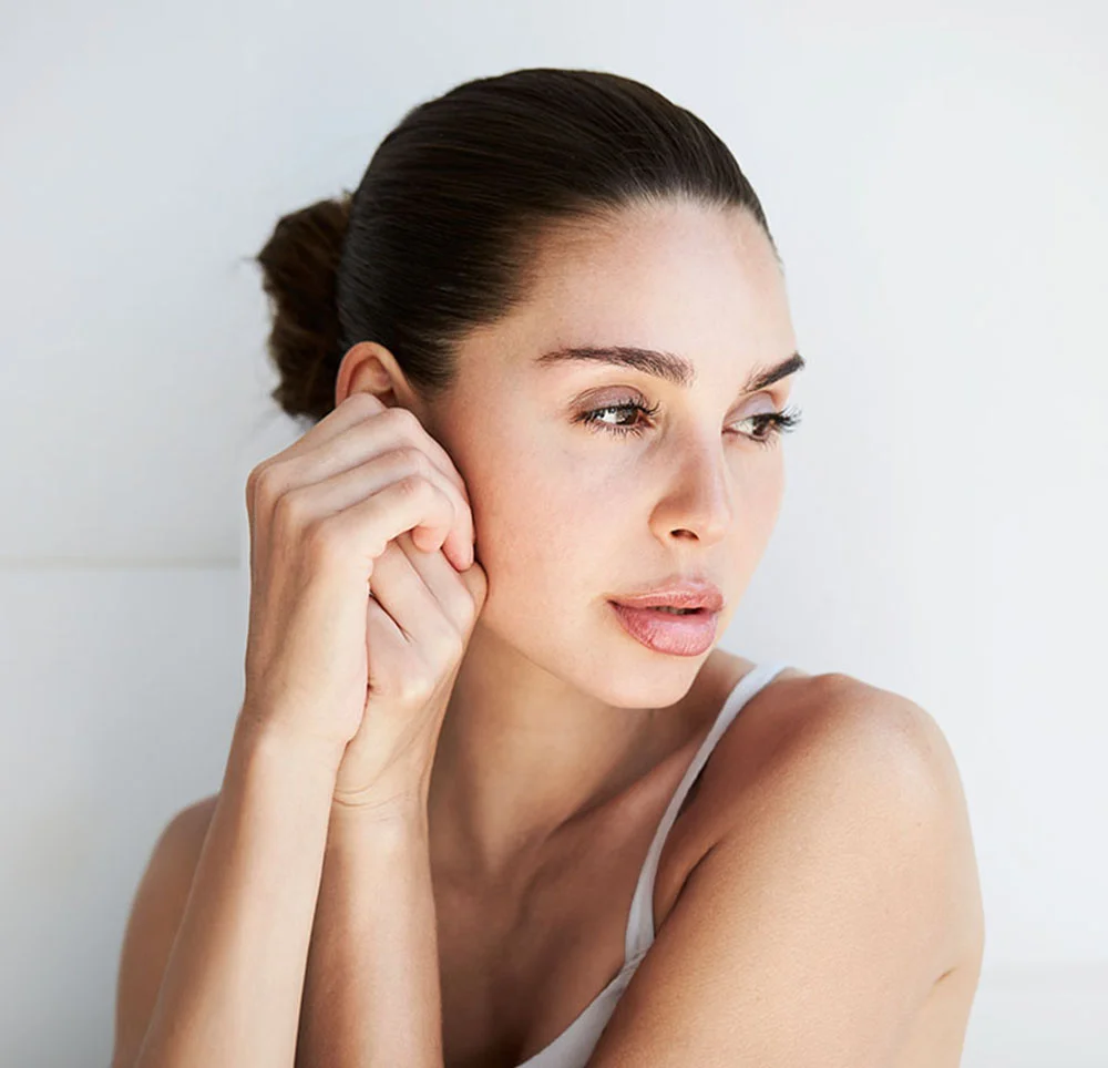 A woman with her brunette hair slicked back in a bun, wearing a white tank top, looking to the side as she holds her hands close to her face - Hand Rejuvenation in Toronto, ON