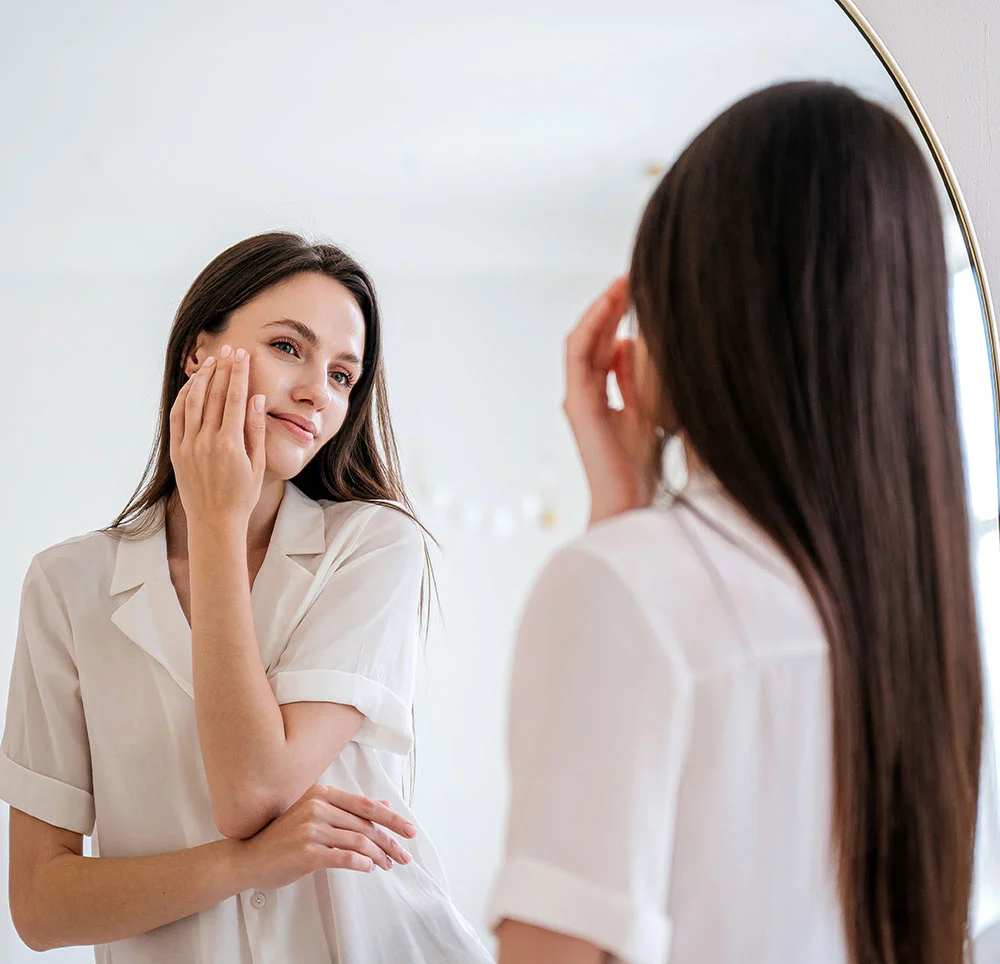 A brunette woman wearing a white button up pajama top, touching her face and looking in her reflection in a large mirror - Microneedling in Toronto, ON
